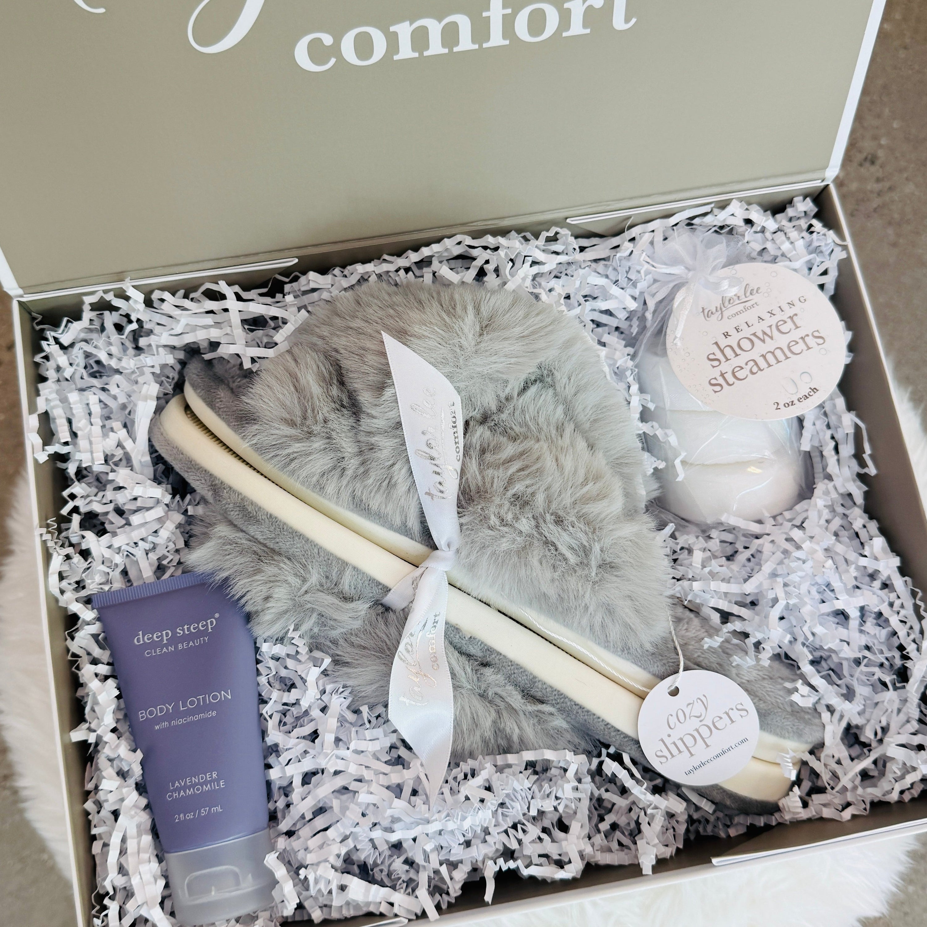 An open gift box labeled Taylor lee Comfort, filled with white shredded paper and containing a gray fluffy slippers, a purple body lotion tube, a small bag of shower steamers. The box lid shows the brand name Taylor lee comfort in white script.
