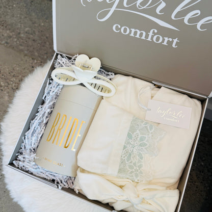 An open gift box from Taylor Lee Comfort containing a cream-colored robe with a lace detail, a wine glass labeled &