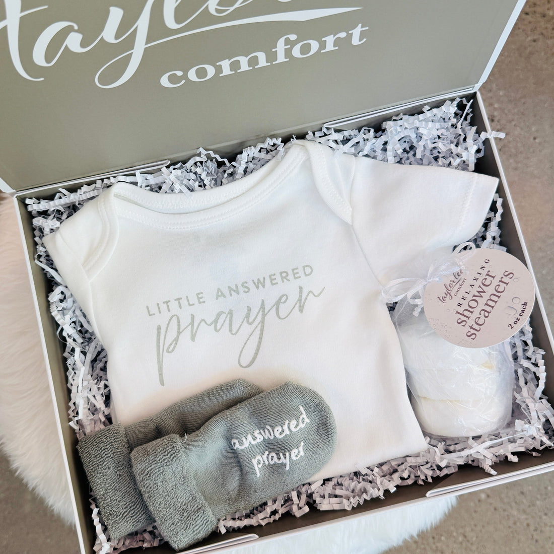 A baby gift set in an open box from Taylor Lee Comfort, featuring a white short-sleeve baby bodysuit with &