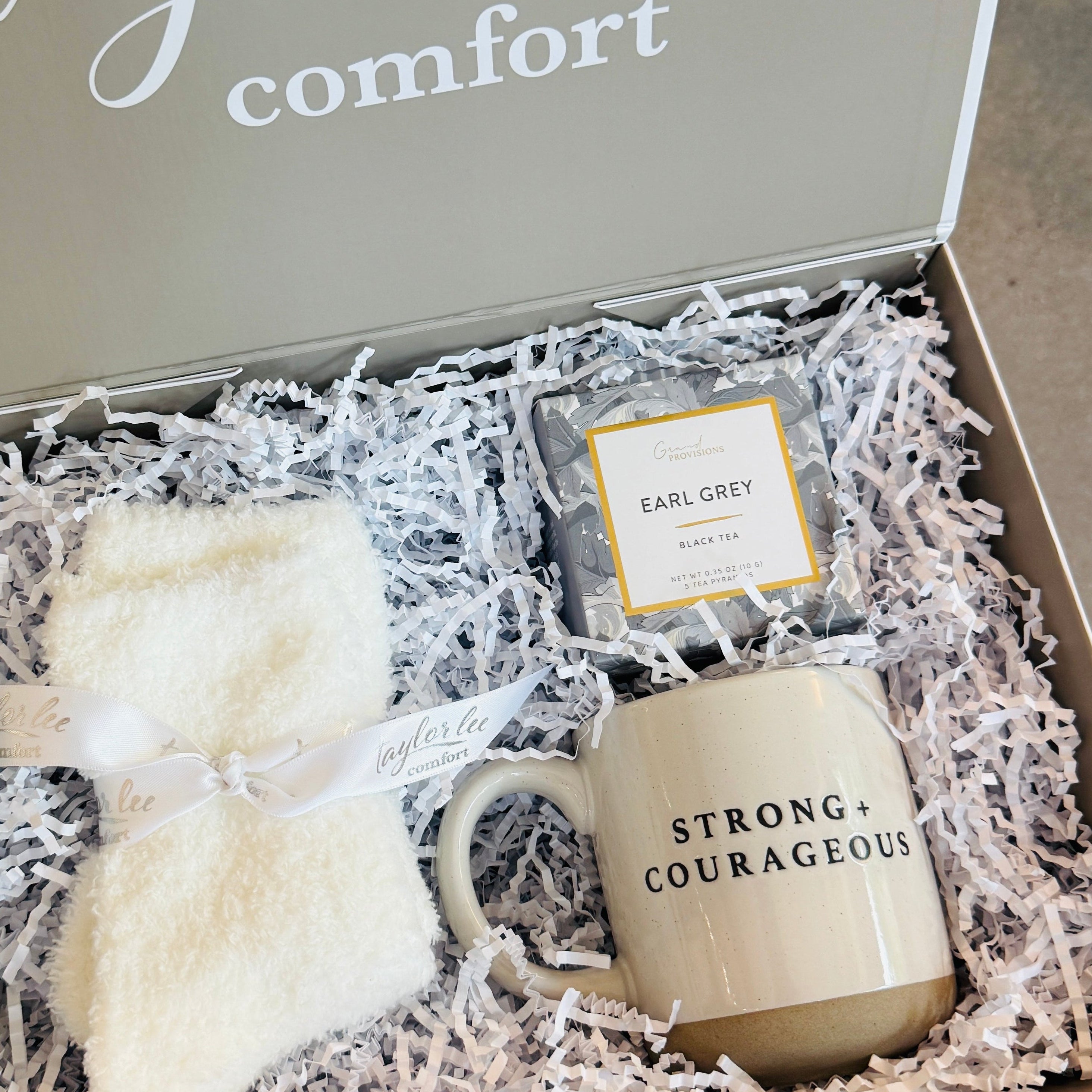 An open gift box from Taylor Lee Comfort sits on a white faux-fur stool. The box is lined with white shredded paper and contains a cream-colored pair of socks tied with a white ribbon, a small package of comforting tea, and a ceramic mug that reads &