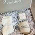 An open gift box from Taylor Lee Comfort sits on a white faux-fur stool. The box is lined with white shredded paper and contains a cream-colored pair of socks tied with a white ribbon, a small package of comforting tea, and a ceramic mug that reads &