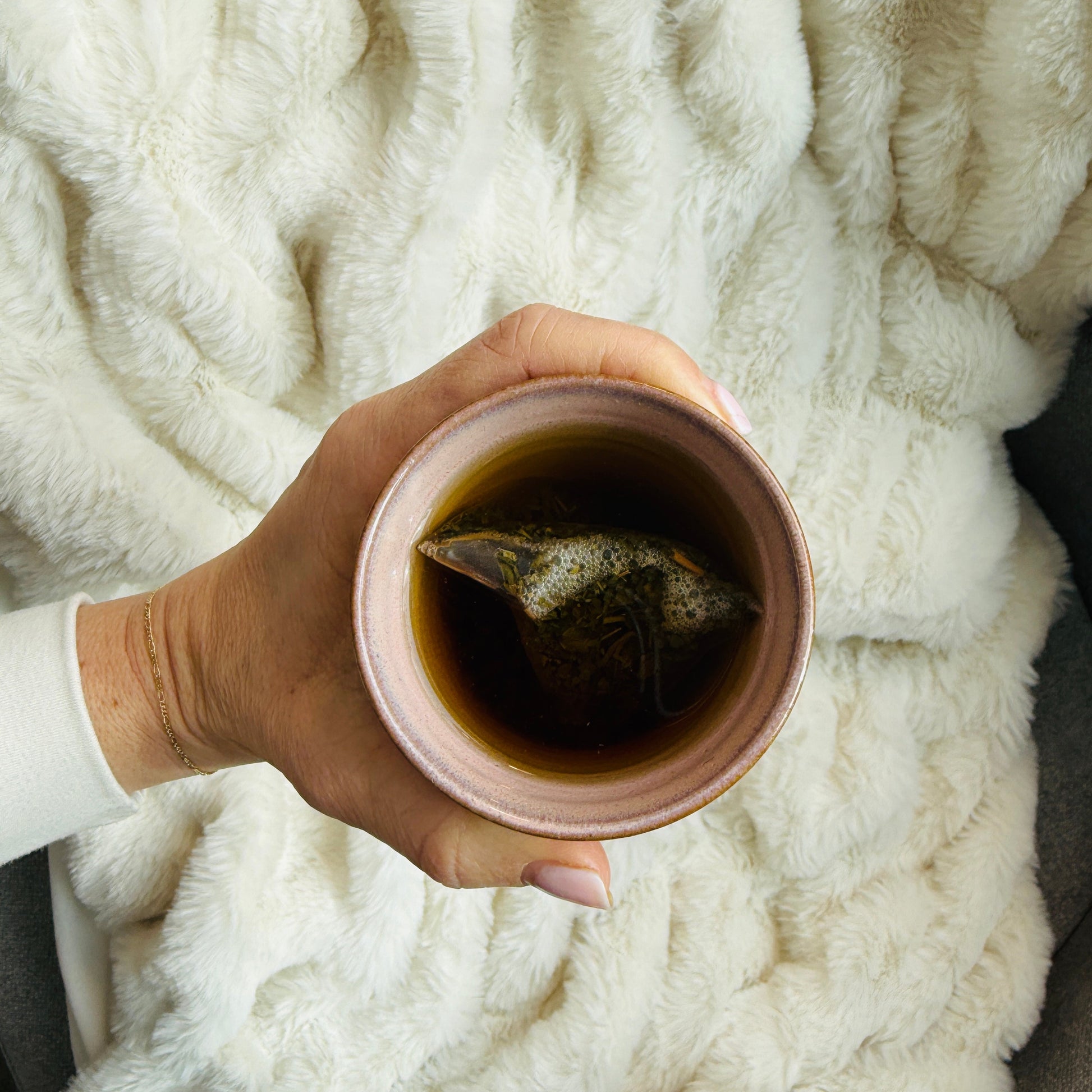 Hand holding a cup of tea with the tea bag inside, wrapped in a fluffy white blanket.