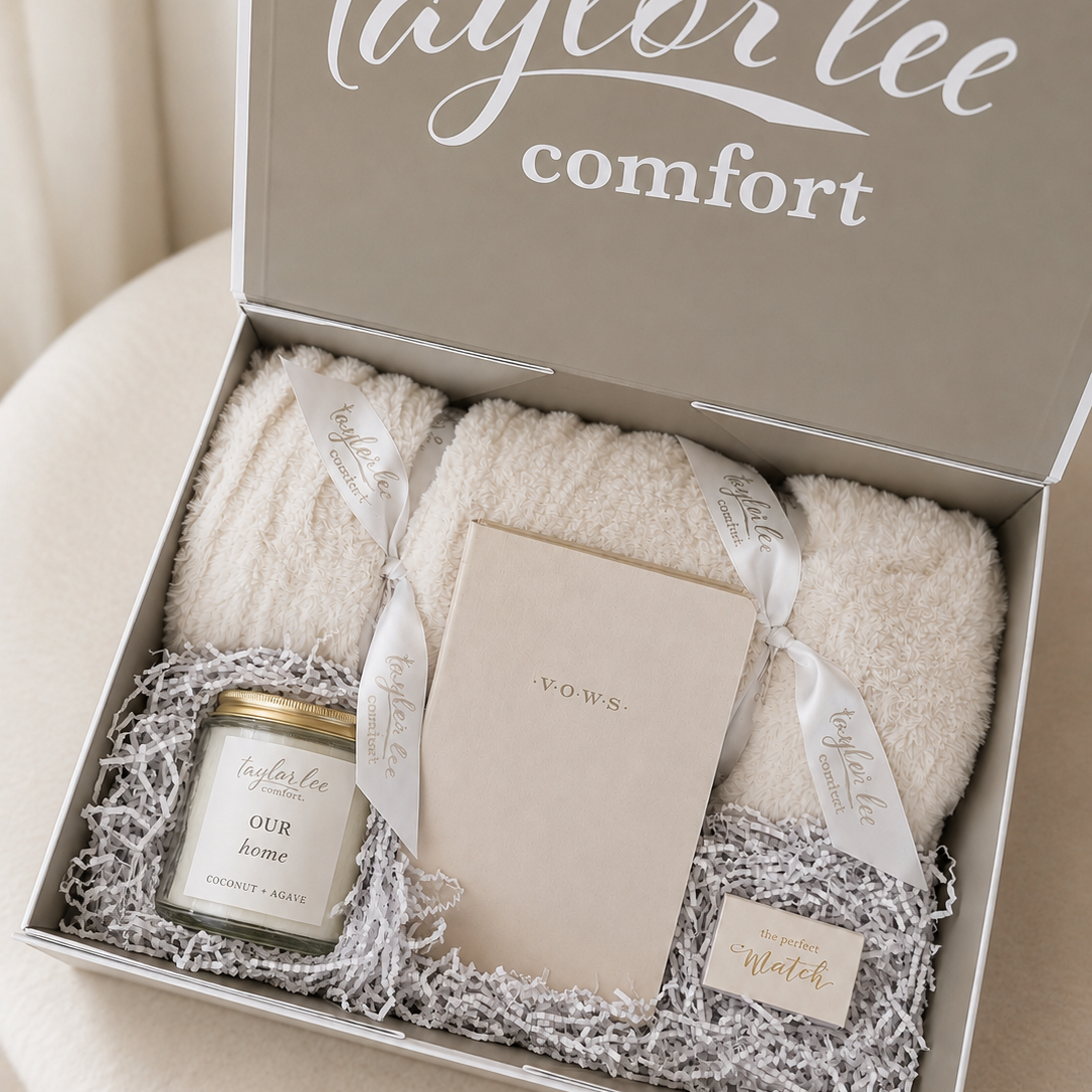 Taylor Lee Comfort wedding or anniversary gift set in an open gift box, featuring cream colored plush blanket, a candle jar, and cream colored journal that says "vows" on the front and safety matches that say "the perfect match" nestled in shredded paper, with the brand name printed on the inside lid.
