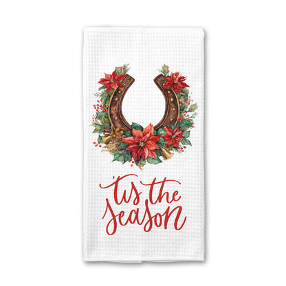A white waffle-weave kitchen towel featuring a festive graphic with a brown horseshoe wreath decorated with red poinsettias and green leaves, above the red cursive text &